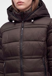 Personne portant une veste noire matelassée à capuche avec fermeture éclair avant et poches zippées, avec le logo Michael Kors sur les fermetures et les trims.