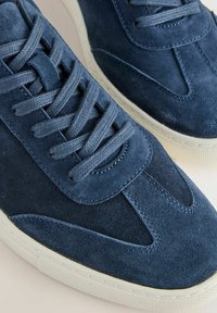 Blå ruskinds sneakers har en rund tå, struktureret overflade og flade snørebånd. Sålen er hvid og let struktureret for bedre greb.