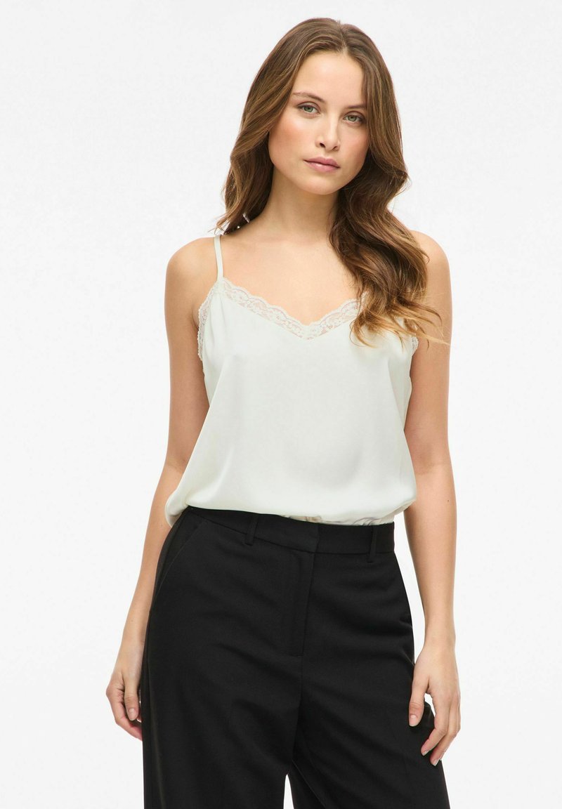 Camisole vert clair avec bordure en dentelle le long du décolleté, fines bretelles et coupe ample, associée à un pantalon noir taille haute.