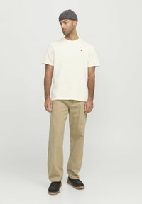 Creme t-shirt med en lille rød detalje, parret med beige vide benbukser og sort fodtøj. Modellen har en grå strikket beanie på.