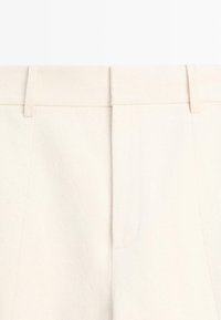 Gros plan sur un pantalon beige taille haute avec passants de ceinture et fermeture éclair frontale dissimulée dans un tissu doux.