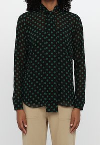 Blouse noire transparente à pois verts avec un nœud au cou, portée avec un pantalon beige.