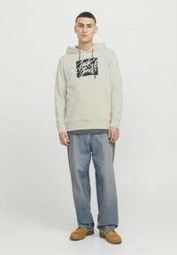 Lys beige hoodie med et sort grafisk logo på forsiden, snore i hætten og en kængurulomme. Båret med afslappede blå jeans og brune sko.