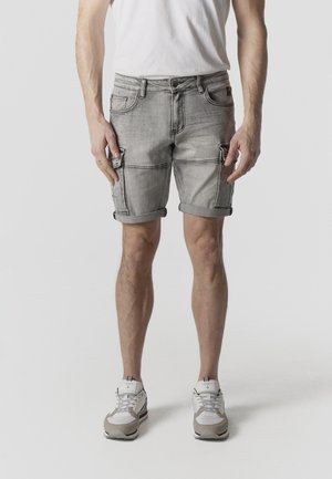 Hombre de pie con pantalones cortos de mezclilla gris claro remangados, zapatillas blancas y camiseta blanca, recortado a la altura de la cintura