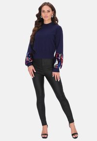 Tengerészkék pulóver, flitteres mintás ujjal, fényes fekete leggingsekkel párosítva, fekete magassarkú szandálokkal viselve. Sima textúra és testhezálló fazon.