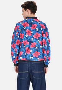 Bloemige bomberjack met een blauwe achtergrond, featuring grote rode hibiscus- en witte bloemen, ribbelkraag en -zoom in zwart, en een gladde textuur.