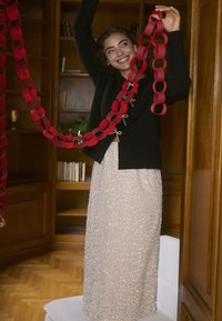 Fekete pulóver masnikkal és egy flitteres bézs szoknya. A modell piros papírláncot tart. Fa könyvespolc és világos fa padló látható.