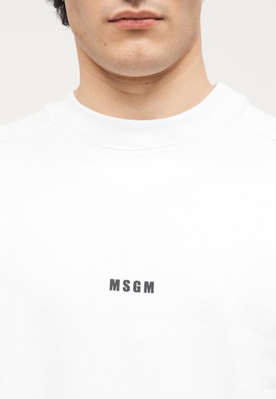 Gros plan sur la partie inférieure du visage et le cou d'une personne portant une chemise blanche avec le texte noir "MSGM" centré sur la poitrine.