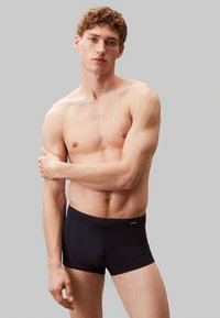 Ciemnoniebieskie bokserki o gładkiej fakturze z metką logo przy pasie, noszone przez modela z kręconymi włosami na neutralnym tle.