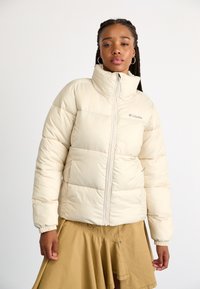 Jachetă puffer bej cu guler înalt și fermoar pe față. Prezintă secțiuni matlasate și manșete elasticizate. Logo-ul Columbia pe piept.