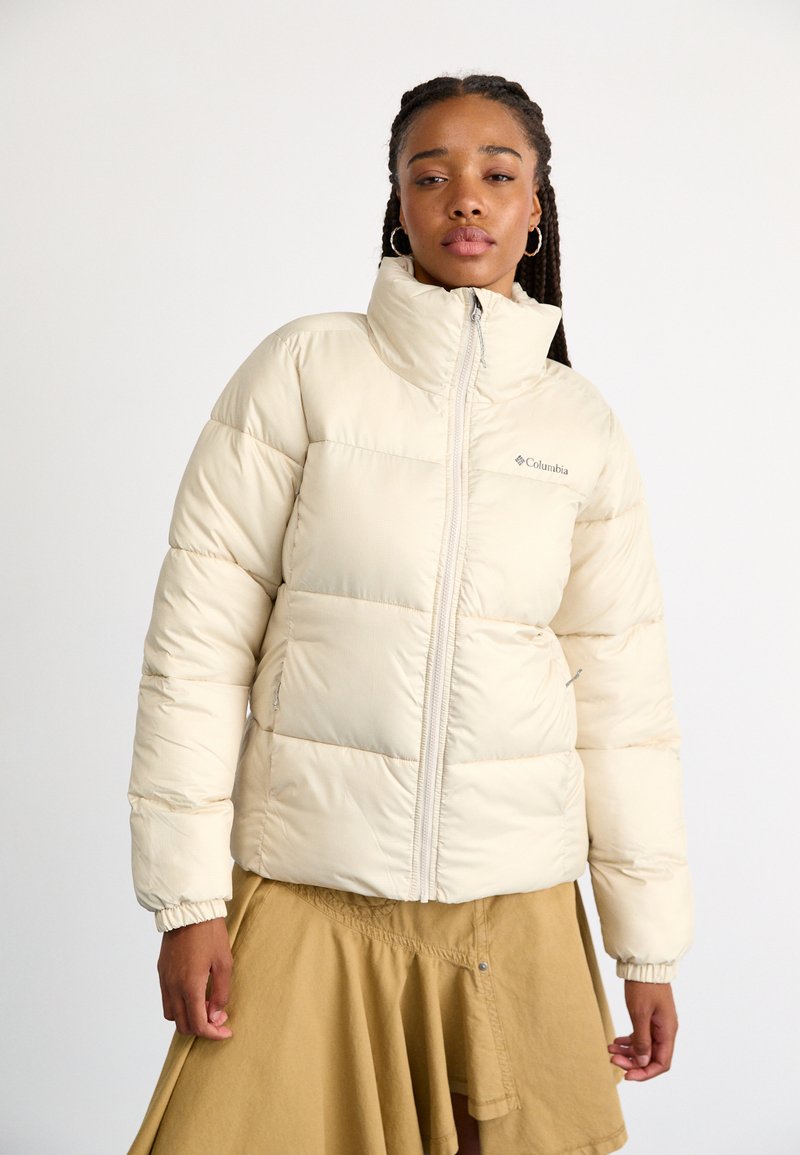 Jachetă puffer bej cu guler înalt și fermoar pe față. Prezintă secțiuni matlasate și manșete elasticizate. Logo-ul Columbia pe piept.