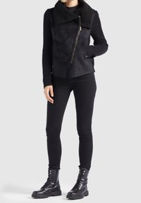 Personne portant un pantalon noir ajusté, des manches en tricot noir, une veste noire asymétrique zippée avec un grand col, et des bottes noires à lacets.