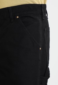 Zwarte denim broek met gouden knoopaccent, voorzien van zijzakken en een gestructureerde stof. Close-up weergave die de stiksels en details benadrukt.