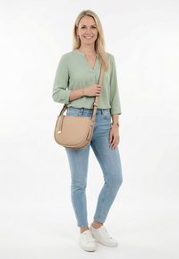 Lächelnde Frau in hellgrüner Bluse, hellblauen Jeans, weißen Turnschuhen und beiger Umhängetasche steht vor weißem Hintergrund.