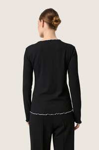 Pruunide juustega naine, kellel on bun soeng, kannab musta tekstuuriga pikkade varrukatega topsi valgete äärtega ning musti pükse, nähtuna tagant beezil taustal.