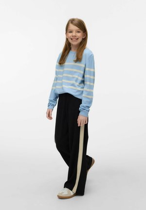 Young girl smiling, wearing a blue and white striped sweater, black wide-leg pants with beige side stripes, and white sneakers.