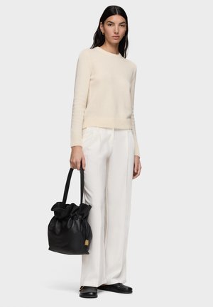Kobieta ubrana w kremowy sweter i białe szerokie spodnie, trzymająca czarną skórzaną torebkę, stojąca na tle jednolitego jasnego tła.