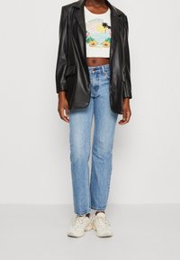 Blazer de imitación de cuero negro sobre una camisa corta blanca con un gráfico colorido de playa, combinado con jeansrectos de mezclilla azul claro y zapatillas beige.