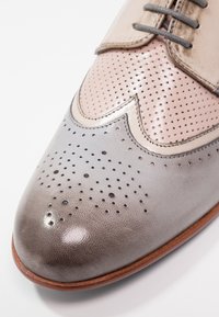 Chaussure brogue en cuir gris avec tige beige perforée, dotée d'un bout pointu et de coutures détaillées, présentant une texture lisse.
