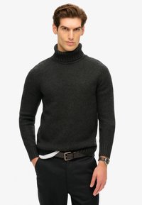 Suéter de cuello alto gris oscuro, con textura acanalada y diseño ajustado. Se lleva con pantalones negros y un cinturón marrón. Acentuado por un reloj en la muñeca.