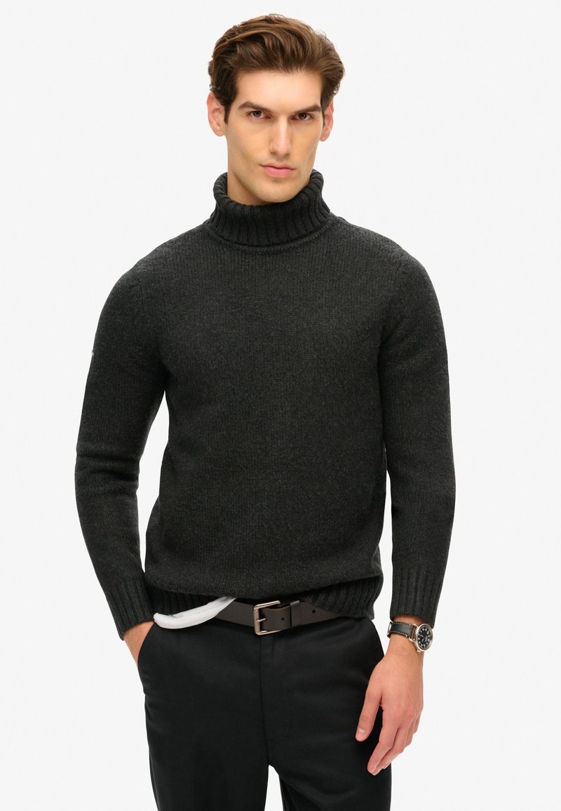Suéter de cuello alto gris oscuro, con textura acanalada y diseño ajustado. Se lleva con pantalones negros y un cinturón marrón. Acentuado por un reloj en la muñeca.