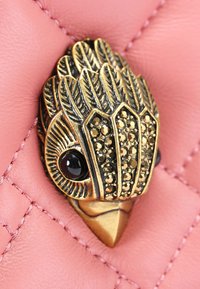 Goudkleurige vogelvormige versiering met donkere edelstenen, geplaatst op een gequilt achtergrond van roze leer met zichtbare stiksels.