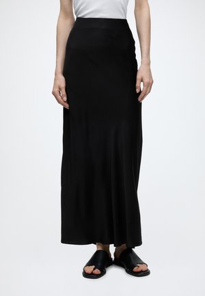 Personne portant une longue jupe noire taille haute, un haut blanc et des sandales noires ouvertes, debout sur un fond blanc uni.