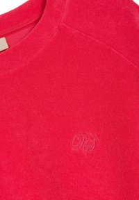 Czerwony sweter z okrągłym dekoltem, miękka tekstura i subtelne haftowane logo w dolnym prawym rogu. Widok z góry, pokazujący wzór i materiał.