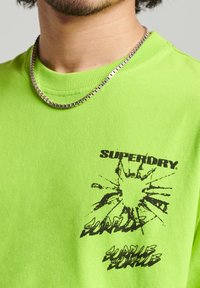 Neon zelené bavlněné tričko s kulatým výstřihem, se černým grafickým designem a textem na levé straně. Viditelný stříbrný řetízek.