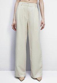 Pantalones de pierna ancha en beis, fabricados en un tejido ligero, con plisados frontales y una textura suave, terminados con bolsillos laterales en ángulo.