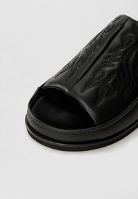 Sandal cu talpă din piele neagră, cu degetul mare descoperit, cusături detaliate în formă de flacără și un finisaj texturat pe materialul superior.