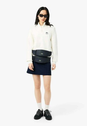 Femme portant un sweat-shirt blanc Lacoste à fermeture éclair quart de zip, une jupe marine, des baskets noires, deux sacs banane noirs, et des lunettes de soleil noires, debout devant un fond blanc.