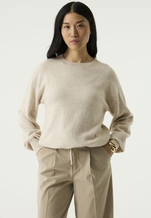 Vrouw met lang donker haar draagt een beige trui en een zandkleurige broek, staand met de handen in de zakken tegen een egale lichte achtergrond.