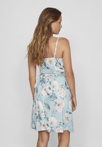 Ziedu kleita ar plāniem siksniņām, gaiši zila auduma, ar lielām rozā un baltām ziedu kompozīcijām, sasieta vidukļa daļā un plūstoša A-formas silueta.