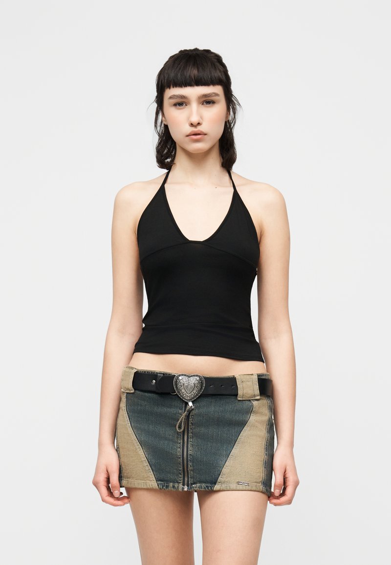 Jeune femme aux cheveux courts et foncés portant un haut noir à col halter et une mini-jupe en denim bicolore avec ceinture à boucle en forme de cœur, debout devant un fond uni.