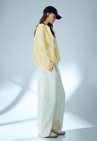 Mlada ženska v ohlapnih belih hlačah, rumeni jakni, črni kapici in belih supergah stoji v minimalističnem studiu z mehko osvetljenimi sencami.