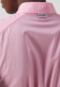 Camicia rosa con colletto classico, texture liscia e leggero lucido; un'etichetta con il marchio "HUGO" è visibile sul retro del collo.