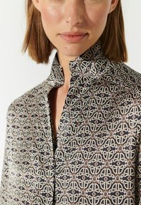 Camicia button-up in tessuto leggero con un motivo geometrico beige e marrone su uno sfondo scuro, con un design del colletto classico.