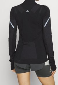 Black athletic long-sleeve top with textured patterns, high collar, and silver accents on sleeves. Features logo at the back and fitted design.