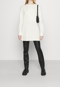 Tunica a maniche lunghe di colore crema con scollo rotondo e un piccolo logo. Abbinata a pantaloni neri aderenti e scarpe nere chunky.