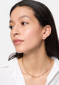 Geometrische Ohrstecker aus Roségold und eine passende Halskette im Papierclip-Stil, getragen mit einem weißen Hemd, das glatte Texturen und einfache Formen zeigt.