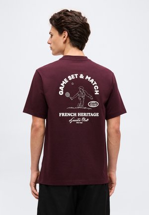 Homme de profil portant un t-shirt bordeaux avec un graphisme blanc sur le thème du tennis et le texte « Game Set & Match » et « French Heritage Lacoste Club ».