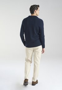 Granatowy sweter z długimi rękawami i kołnierzykiem, zestawiony z jasnobeżowymi spodniami. Całość uzupełniają brązowe buty.