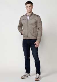 Jachetă din suede gri deschis cu guler în picioare, două buzunare pe piept cu fermoar și accente texturate pe umeri. Pur tată peste o cămașă albă și blugi blue.