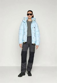 Sieviete, tērpusies gaiši zilā pufīgajā jaku, pelēkā džemperī, melnās cargo biksēs, melnās zālēs un lielās melnās saulesbrillēs, stāv pret tukšu fonu.