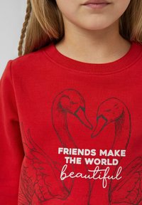 Kind dat een rood T-shirt draagt met twee zwanen en de tekst "Vrienden maken de wereld mooi" op de voorkant gedrukt.
