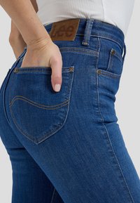 Gros plan d'une personne portant un jean bleu Lee, la main dans la poche arrière, et une chemise blanche rentrée.