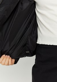 Black jacket with a shiny, smooth interior. Features a drawstring at the hem and a subtle logo inside. Exterior is matte and textured.