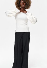 Vrouw met krullend haar die de halslijn van een witte trui met lange mouwen aanpast, gekleed in zwarte wijde pantalon, tegen een witte achtergrond.