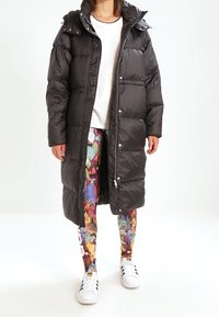 Dlouhý černý péřový kabát se zipem a patentkami, který má kapuci a design s prošitými panely, v kombinaci s barevně vzorovanými legínami a teniskami.